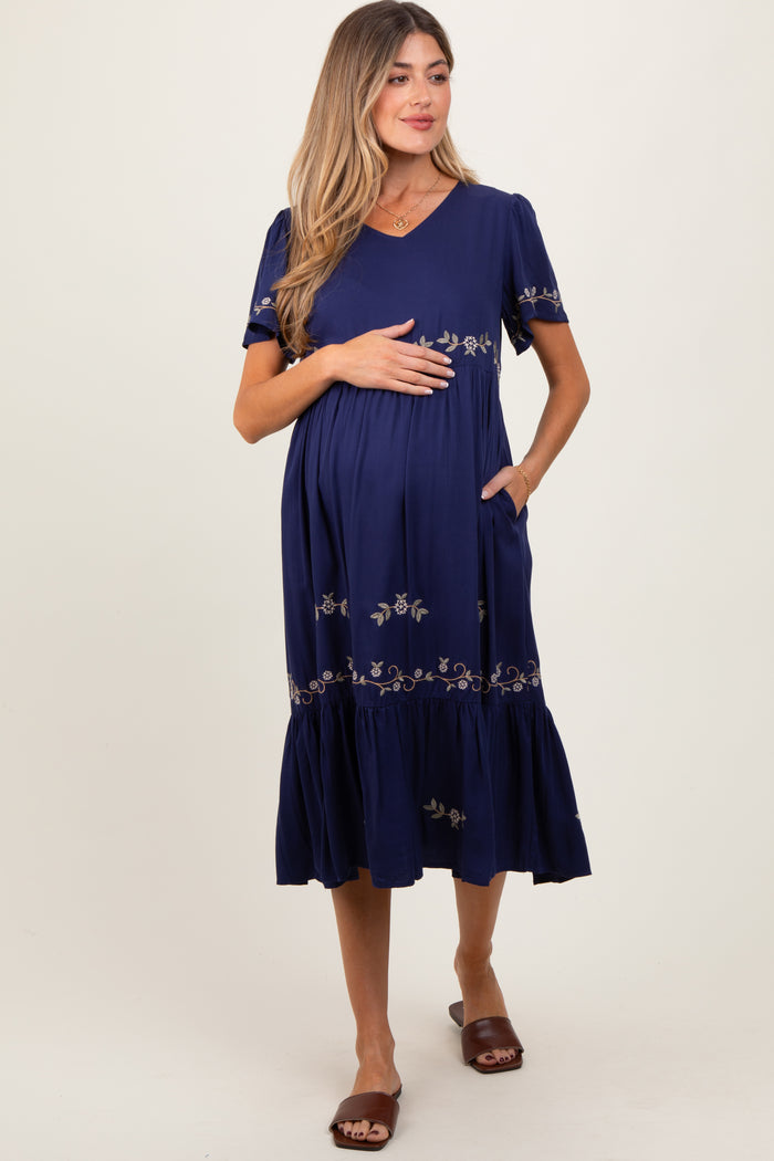 woman wearing a navy blue maternity dress with short sleeves, v-neck, and floral embroidery, highlighting her baby bump