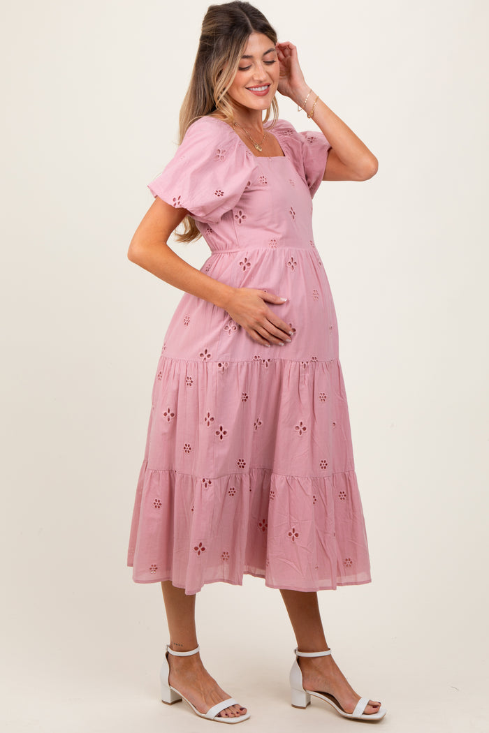 woman wearing a pink maternity dress with puff sleeves and eyelet details, gently cradling her baby bump
