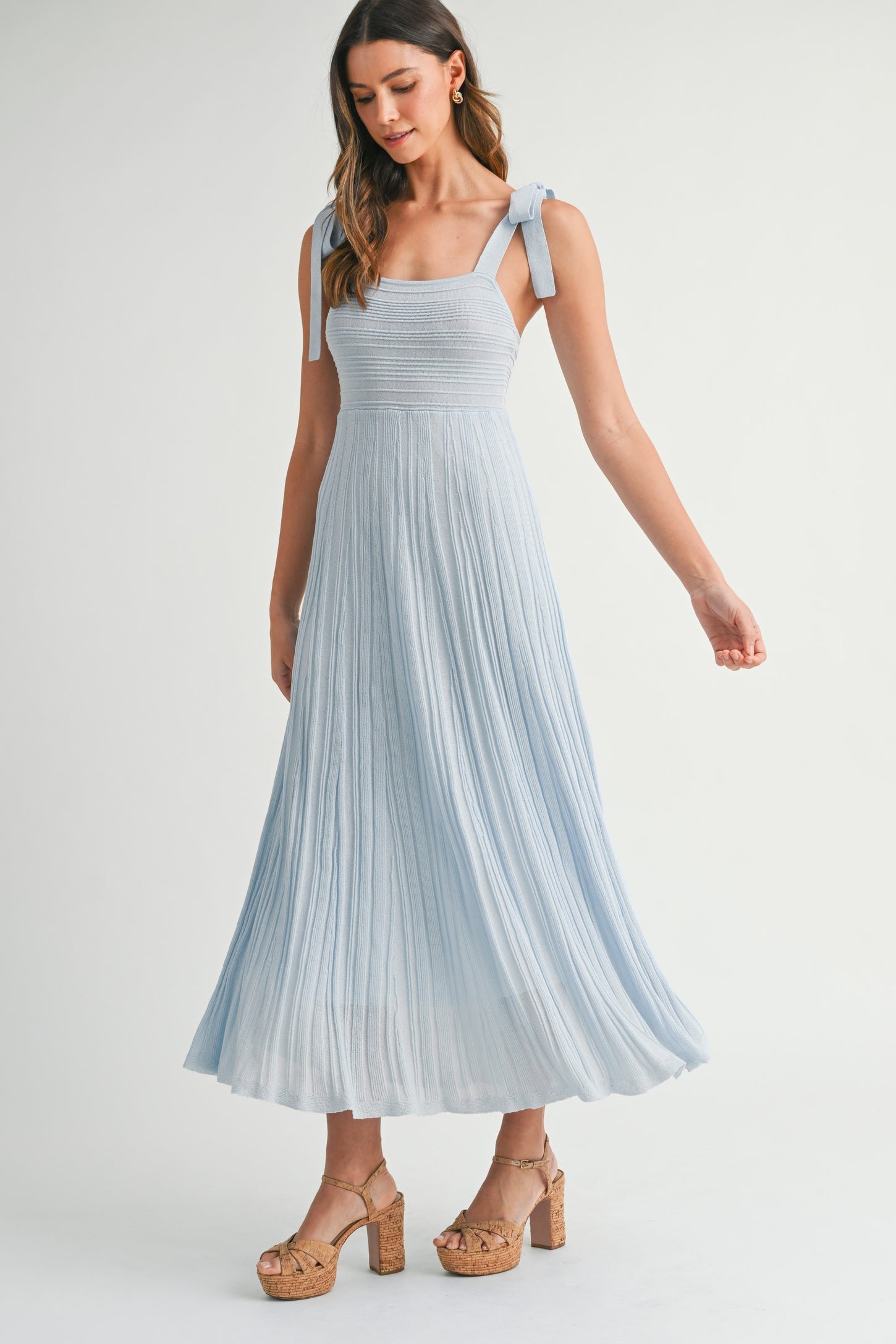 woman wearing a light blue pleated maxi dress with adjustable tie straps and platform wedge heels