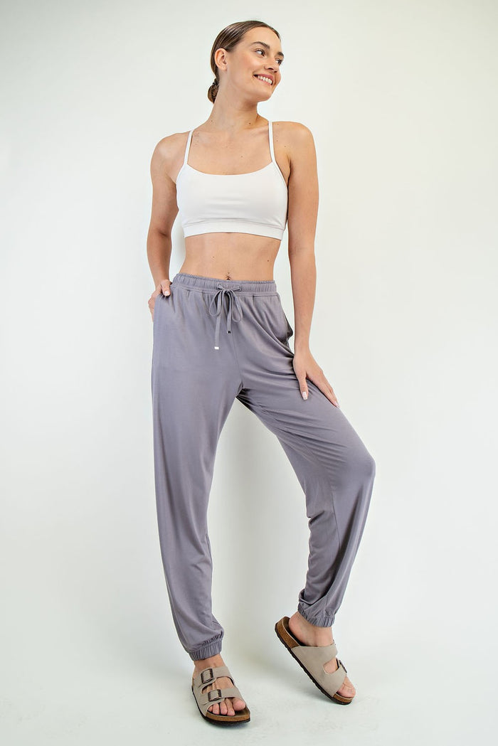 woman wearing a white cropped tank top and light gray jogger pants, standing with hands in pockets and smiling