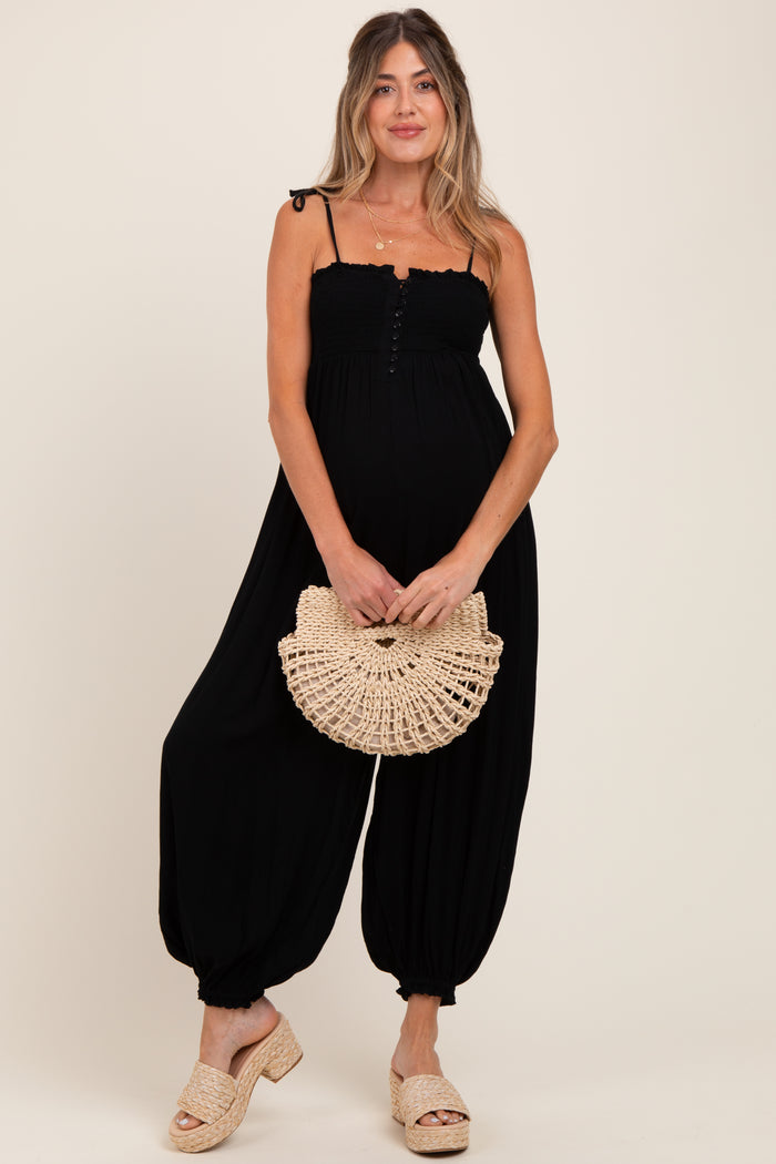 woman wearing a black maternity jumpsuit with smocked bodice and wide legs, holding a straw handbag and showing her baby bump