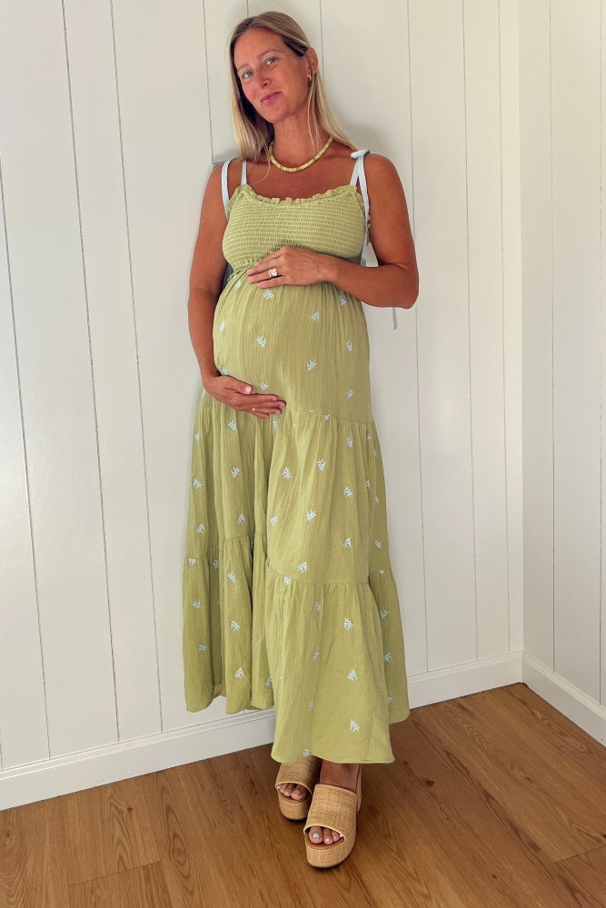 woman wearing a green maternity dress with tiered ruffles and white embroidery, holding her baby bump