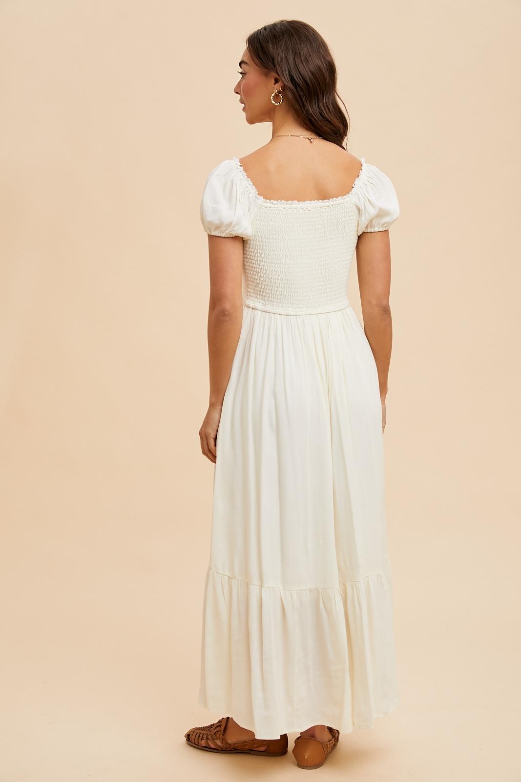 woman wearing a white maxi dress with a smocked bodice and puff sleeves, showing the back view against a beige background