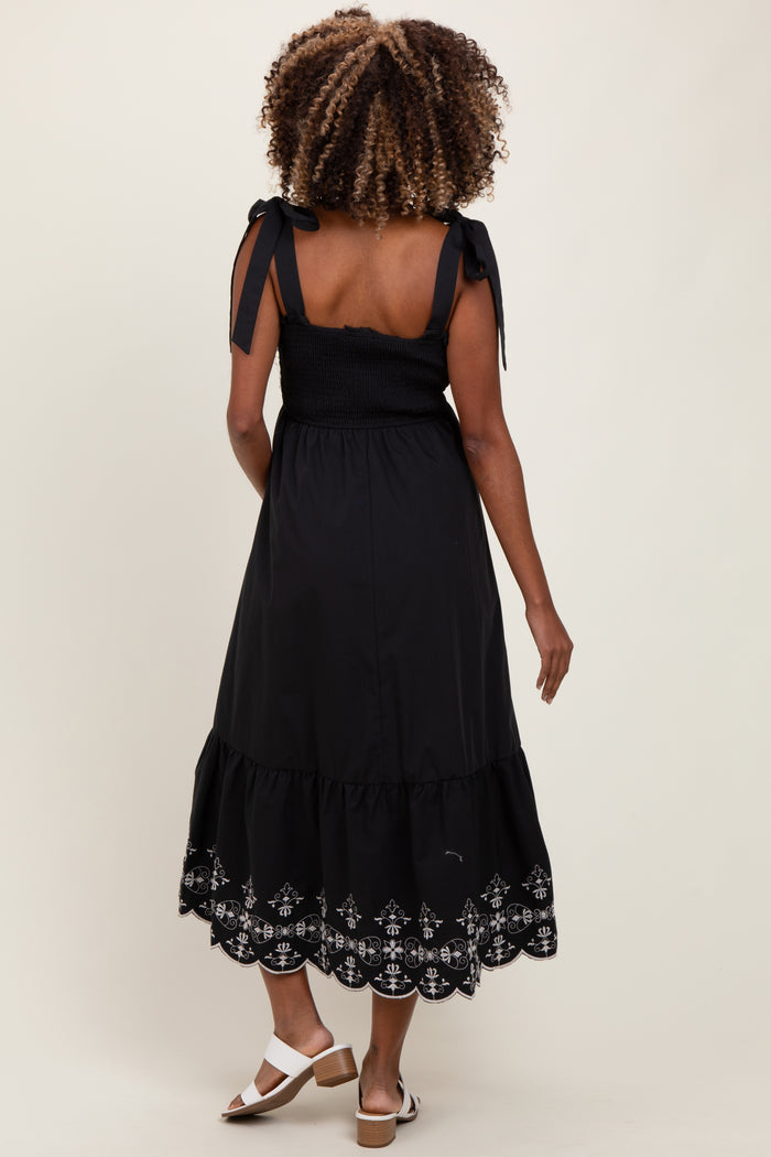 woman wearing a black maternity dress with tie straps and embroidered hem, showcasing a baby bump