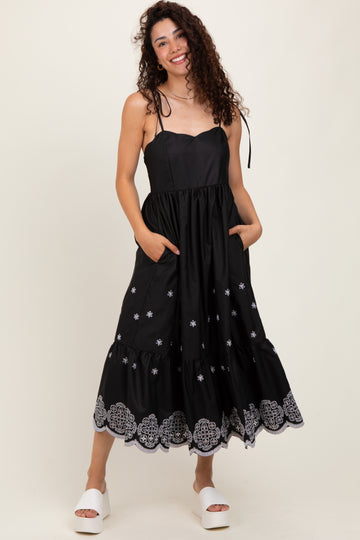 woman wearing a black midi dress with white embroidery and pockets, featuring adjustable straps and a tiered skirt.