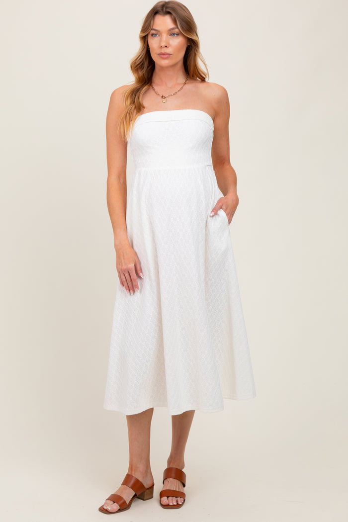 woman wearing a white strapless midi maternity dress with a textured pattern and side pockets, standing on a neutral background