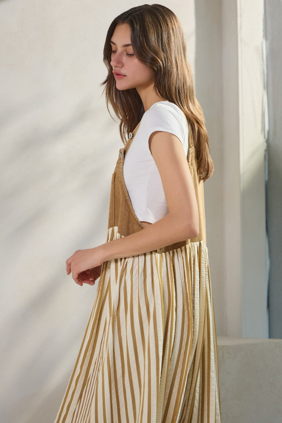 woman wearing a white short sleeve top under a tan striped pinafore dress, standing against a light background
