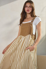 woman wearing a brown corduroy overall dress with striped skirt and white t-shirt underneath, posing with hands in pockets