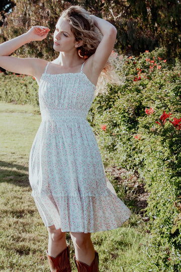 woman wearing a white floral sundress with a ruffled hem and spaghetti straps, posing outdoors among greenery and flowers