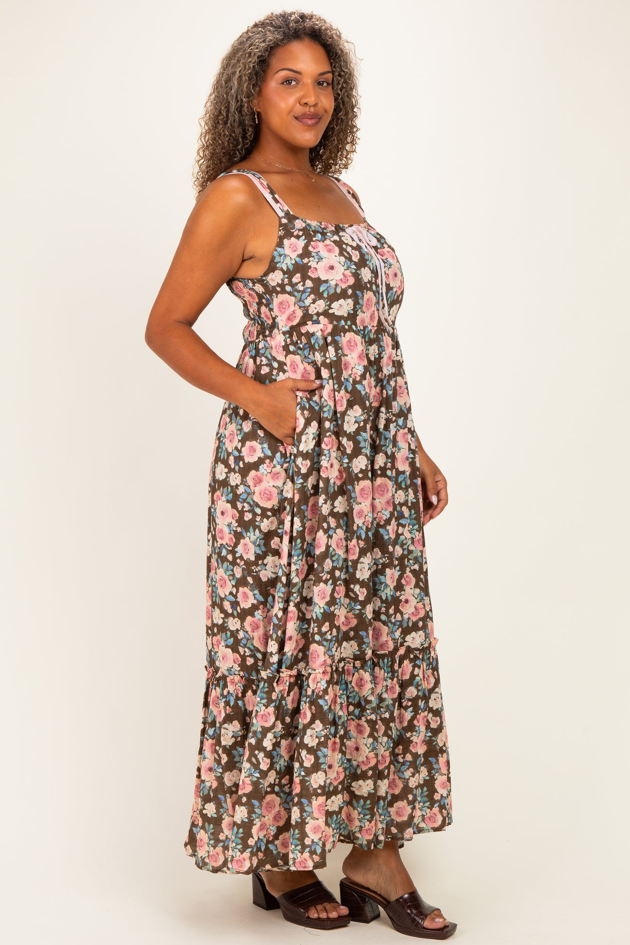 woman wearing a floral maxi dress with pockets in shades of brown, pink, and green, standing against a light background