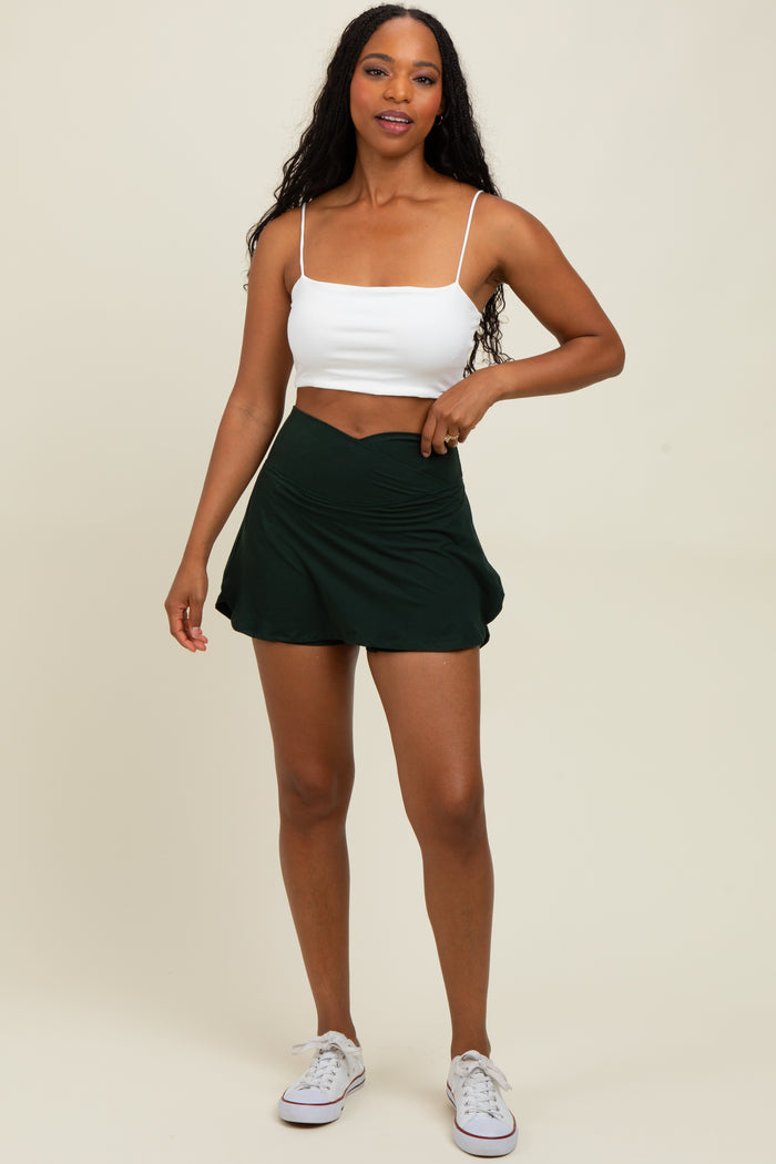 woman wearing a white cropped tank top and a dark green pleated skort, standing with one hand on her hip