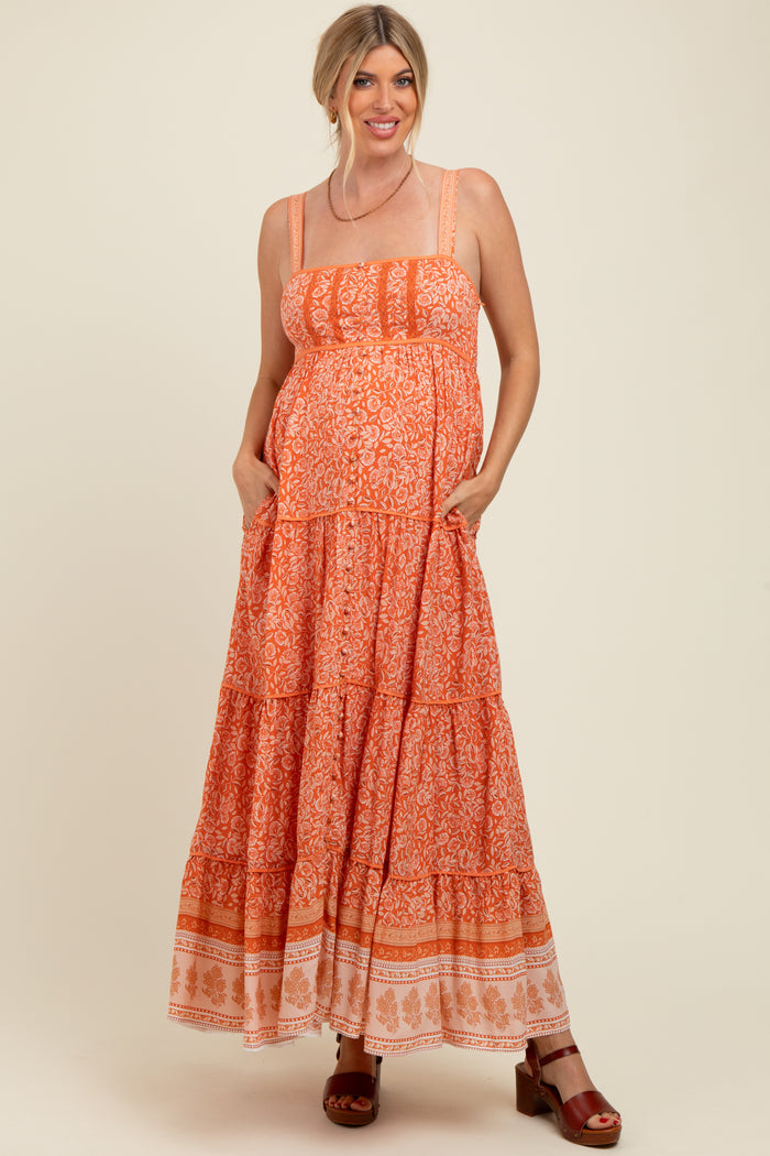 woman wearing an orange patterned maternity maxi dress with tiered details and pockets, smiling while showcasing her baby bump