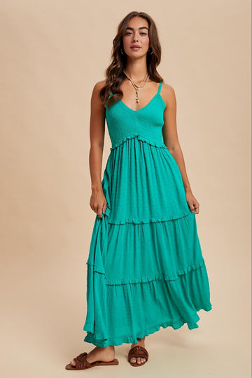 woman wearing a teal tiered maxi dress with thin straps and textured detailing, standing confidently against a neutral background