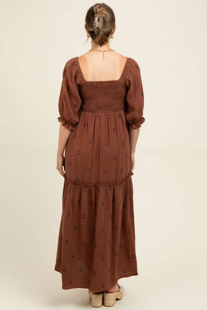 woman wearing a brown maternity maxi dress with puff sleeves and floral embroidery, showing the back view