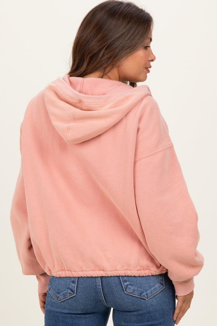 woman wearing a pink maternity hoodie with a drawstring hood and elastic hem, styled with dark jeans