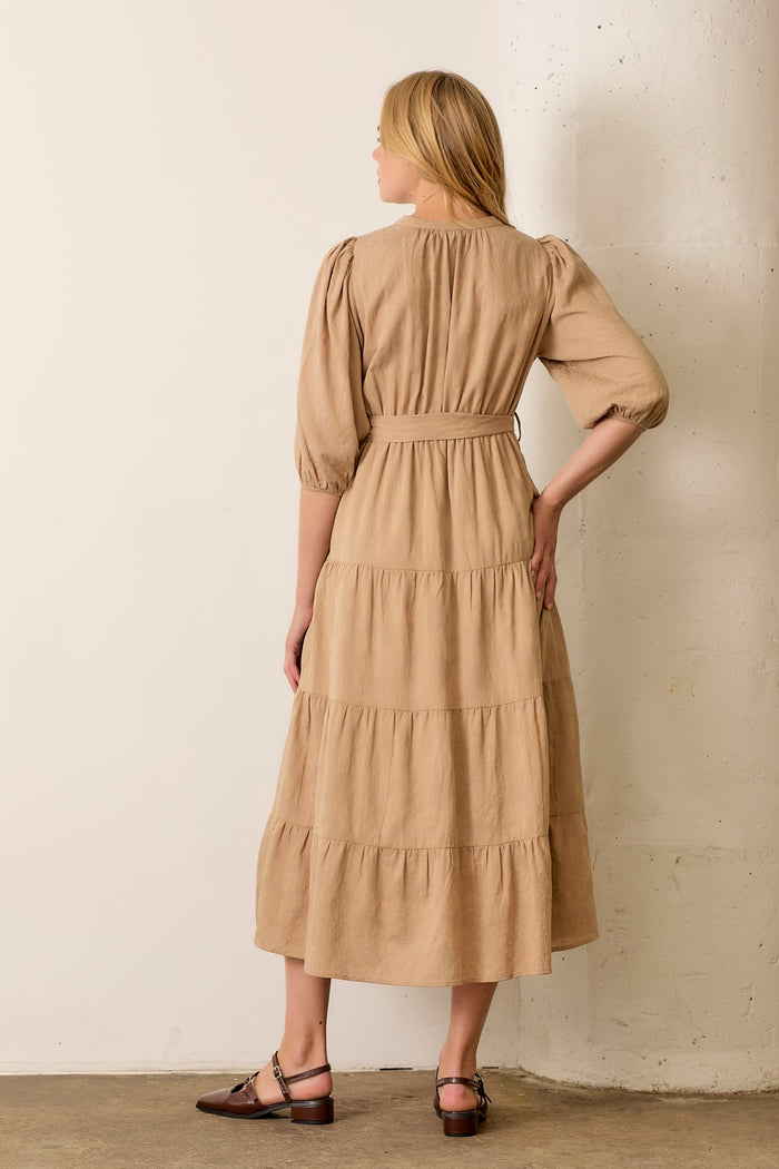 woman wearing a tan tiered midi dress with puff sleeves and a belted waist, standing with her back to the camera
