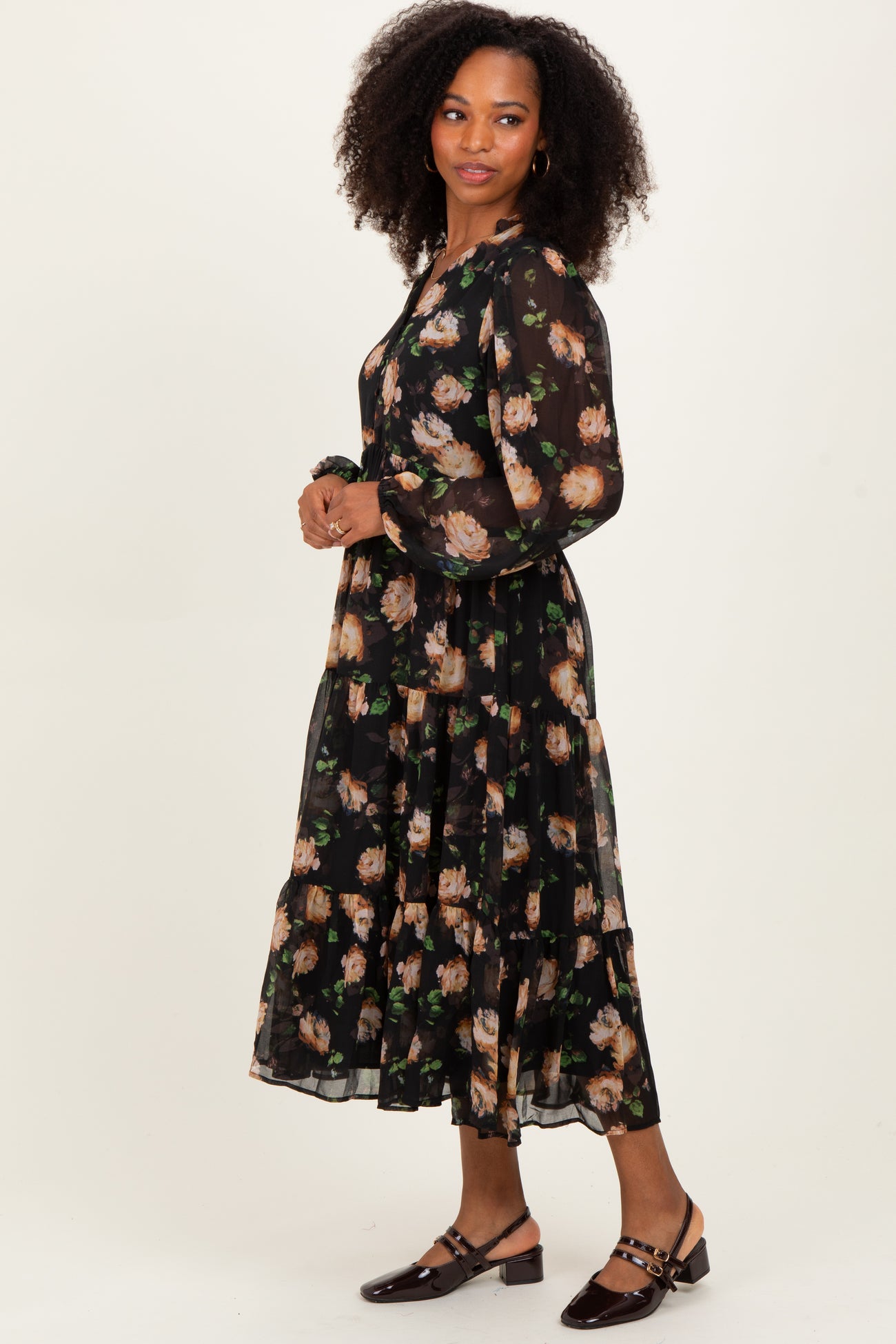 woman wearing a black floral maxi dress with long sleeves and ruffled tiered detailing, standing against a plain background