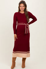 woman wearing a burgundy maternity sweater dress with ribbed texture and a matching tie belt, posing with a smile
