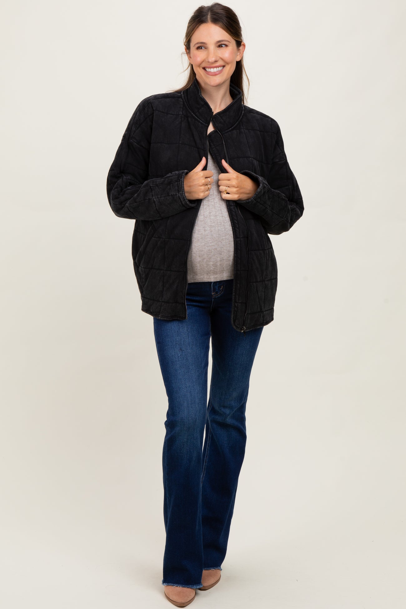 woman wearing a black quilted maternity jacket and a gray top, holding her baby bump while standing in flared jeans