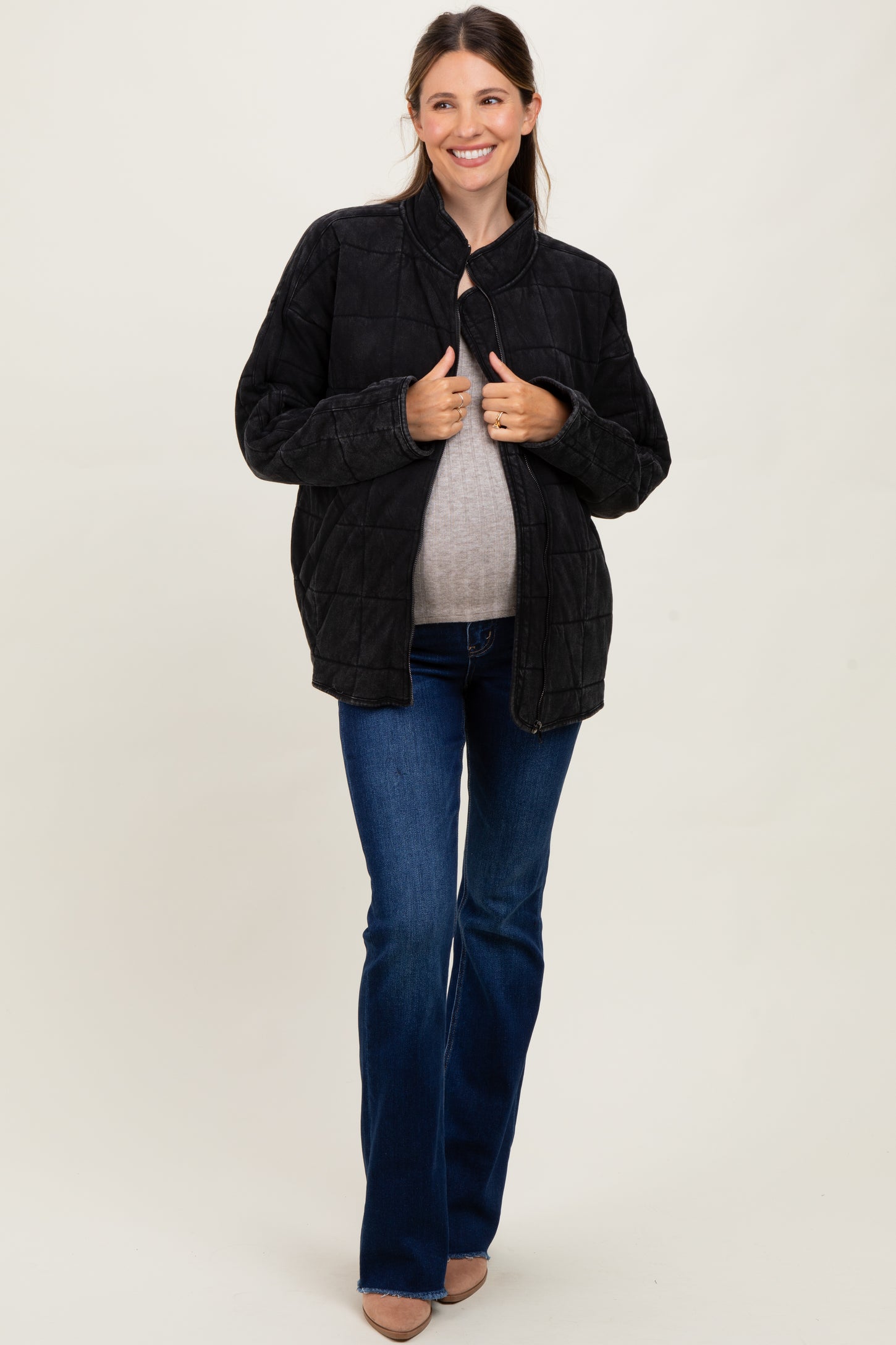 woman wearing a black quilted maternity jacket and a gray top, holding her baby bump while standing in flared jeans