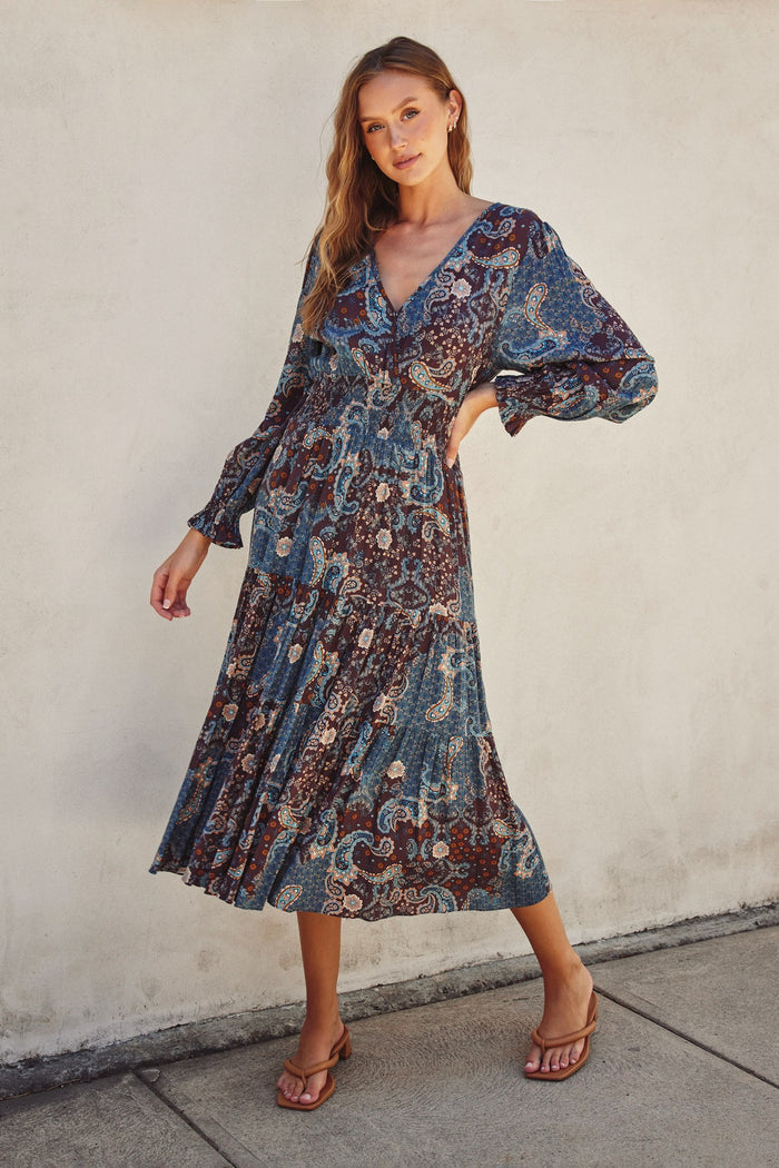 woman wearing a long sleeve paisley print maxi dress with a deep v-neck and tiered skirt, standing on a sidewalk
