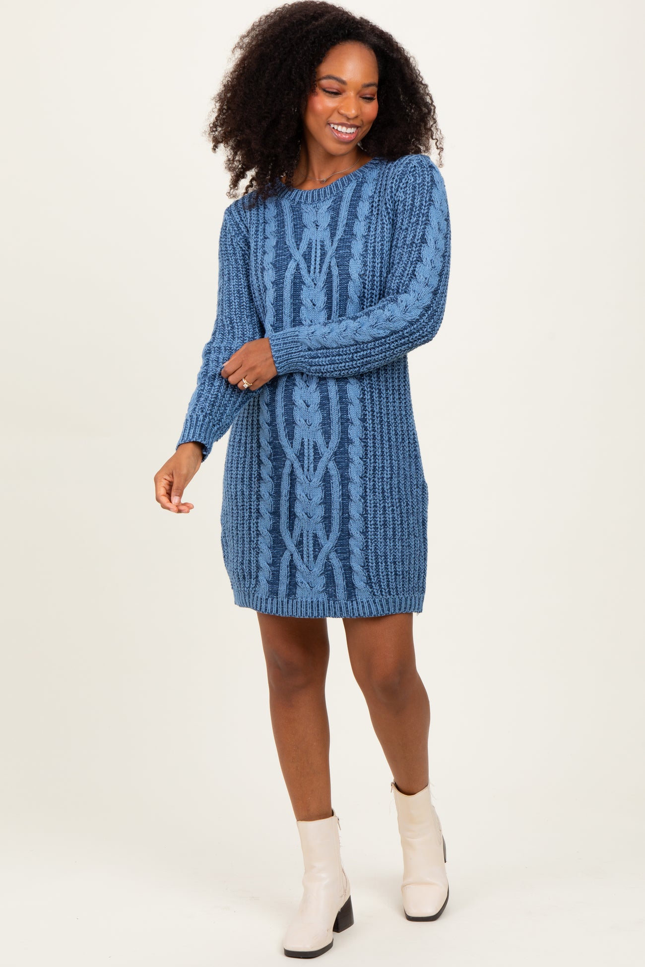 woman wearing a blue cable knit sweater dress and white ankle boots, smiling and posing on a neutral background