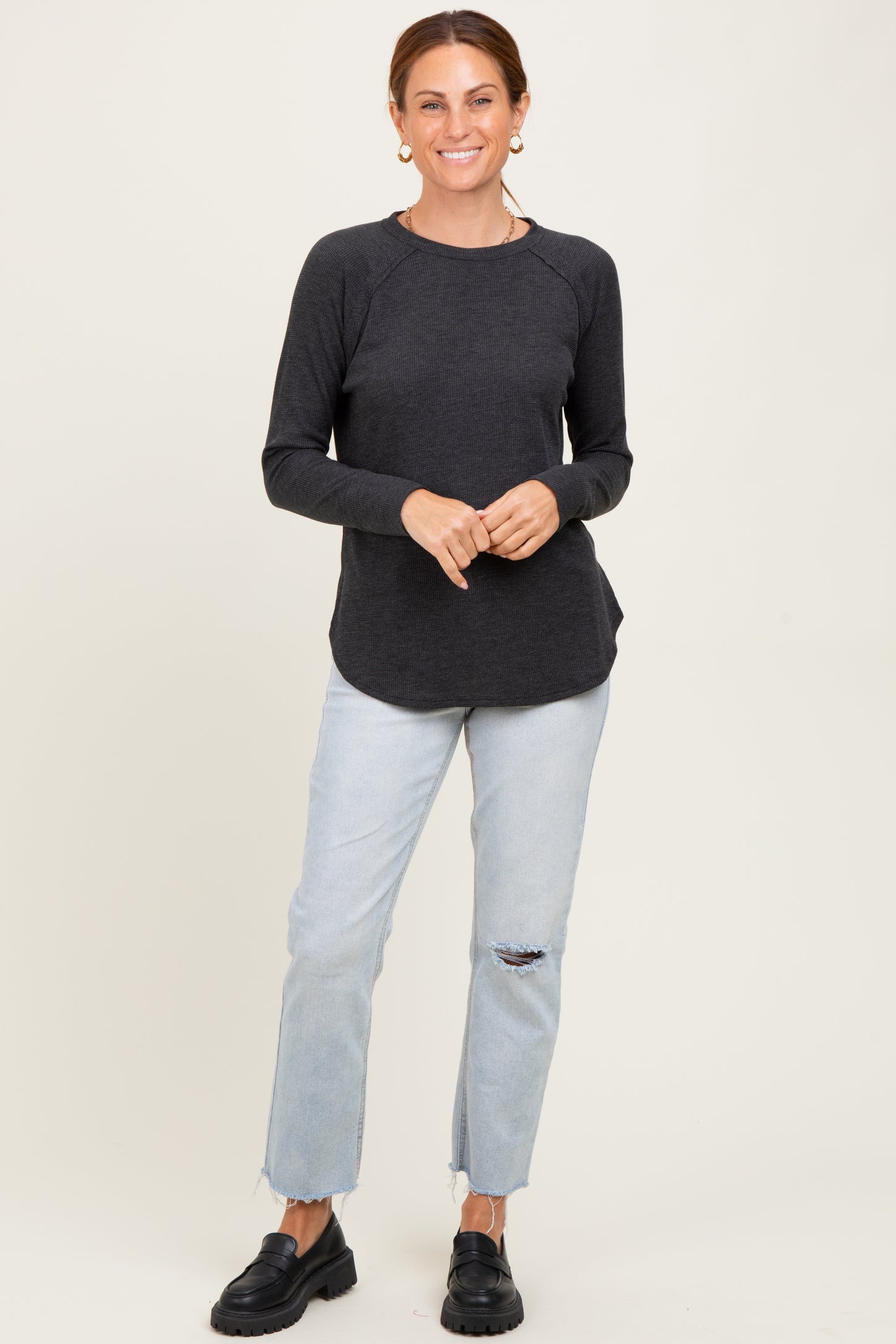 woman wearing a charcoal knit long sleeve top and light blue distressed jeans, standing with hands on hips