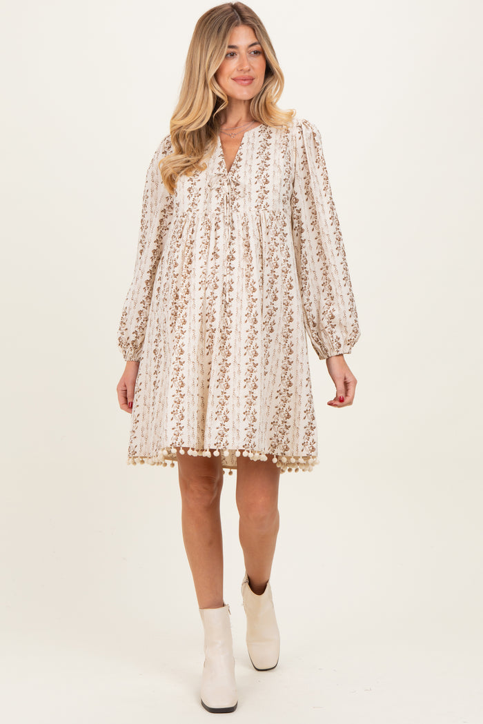 woman wearing a beige floral maternity dress with long sleeves and pom-pom trim, showcasing her baby bump