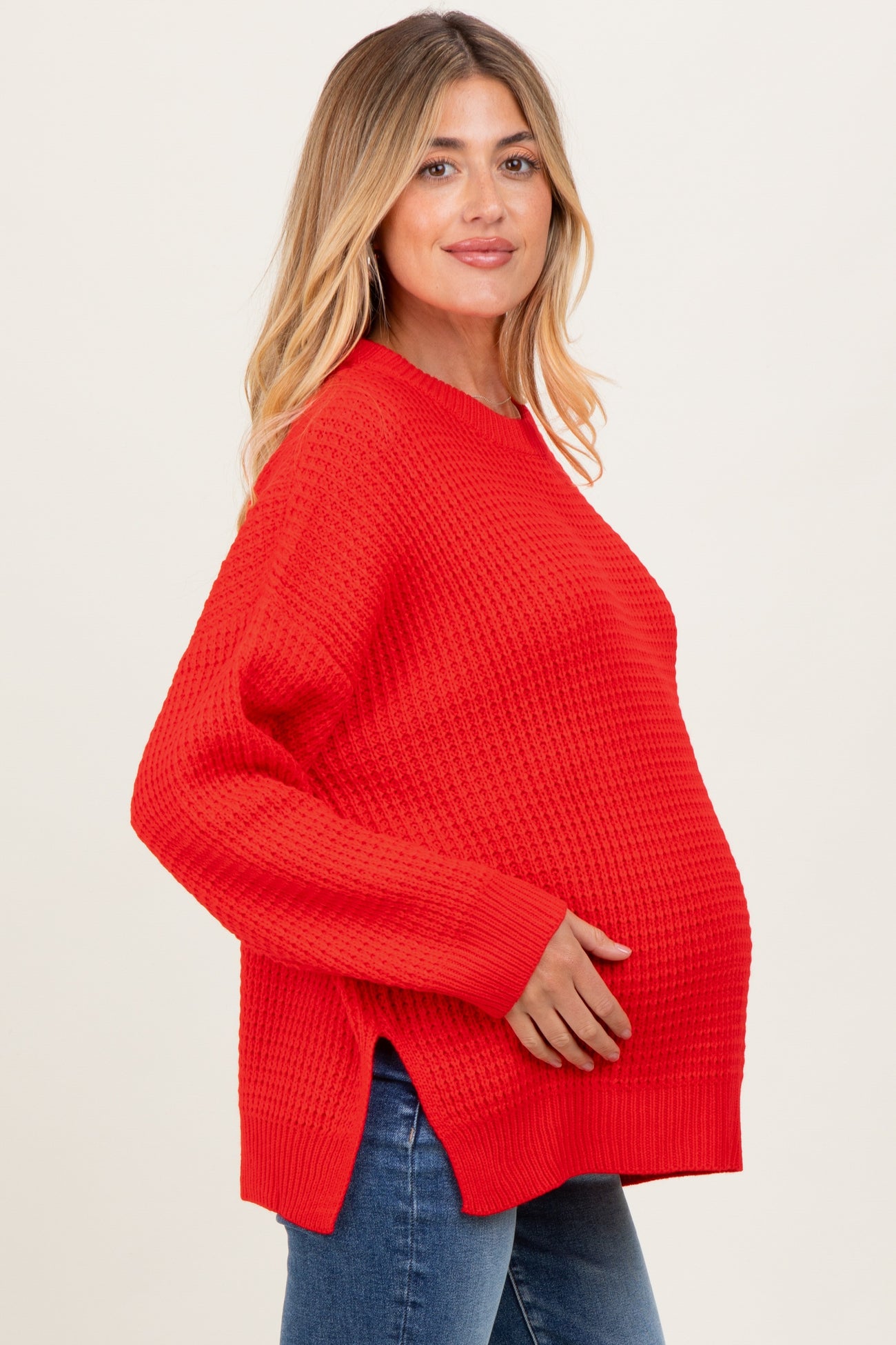 woman wearing a bright red oversized maternity sweater, cradling her baby bump, styled with blue jeans