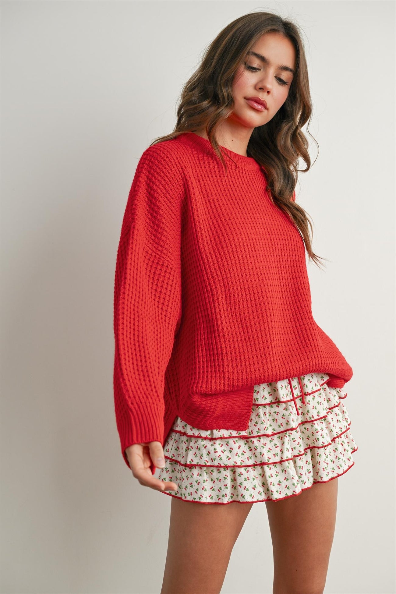 woman wearing a bright red oversized sweater with a textured finish and paired with a white floral ruffled mini skirt
