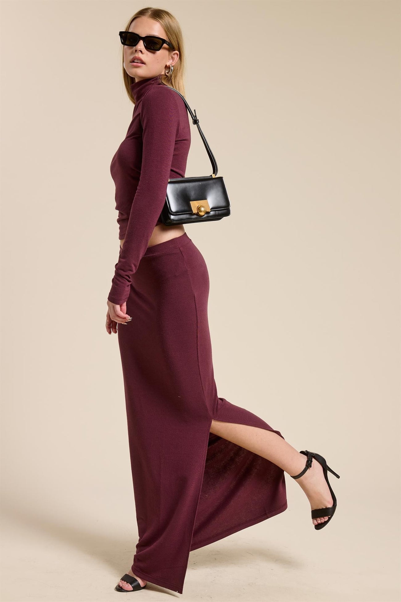 woman wearing a burgundy long sleeve turtleneck top and matching maxi skirt with a high slit, accessorized with a black handbag and sunglasses