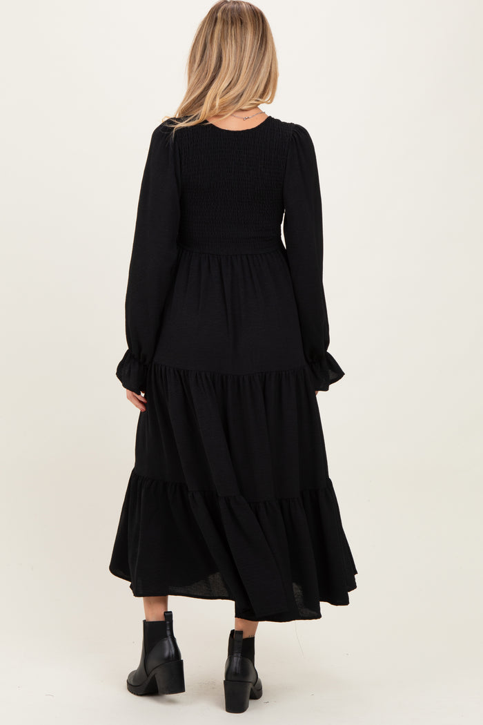 woman wearing a black maternity maxi dress with long sleeves and tiered skirt, showing a smocked bodice from the back