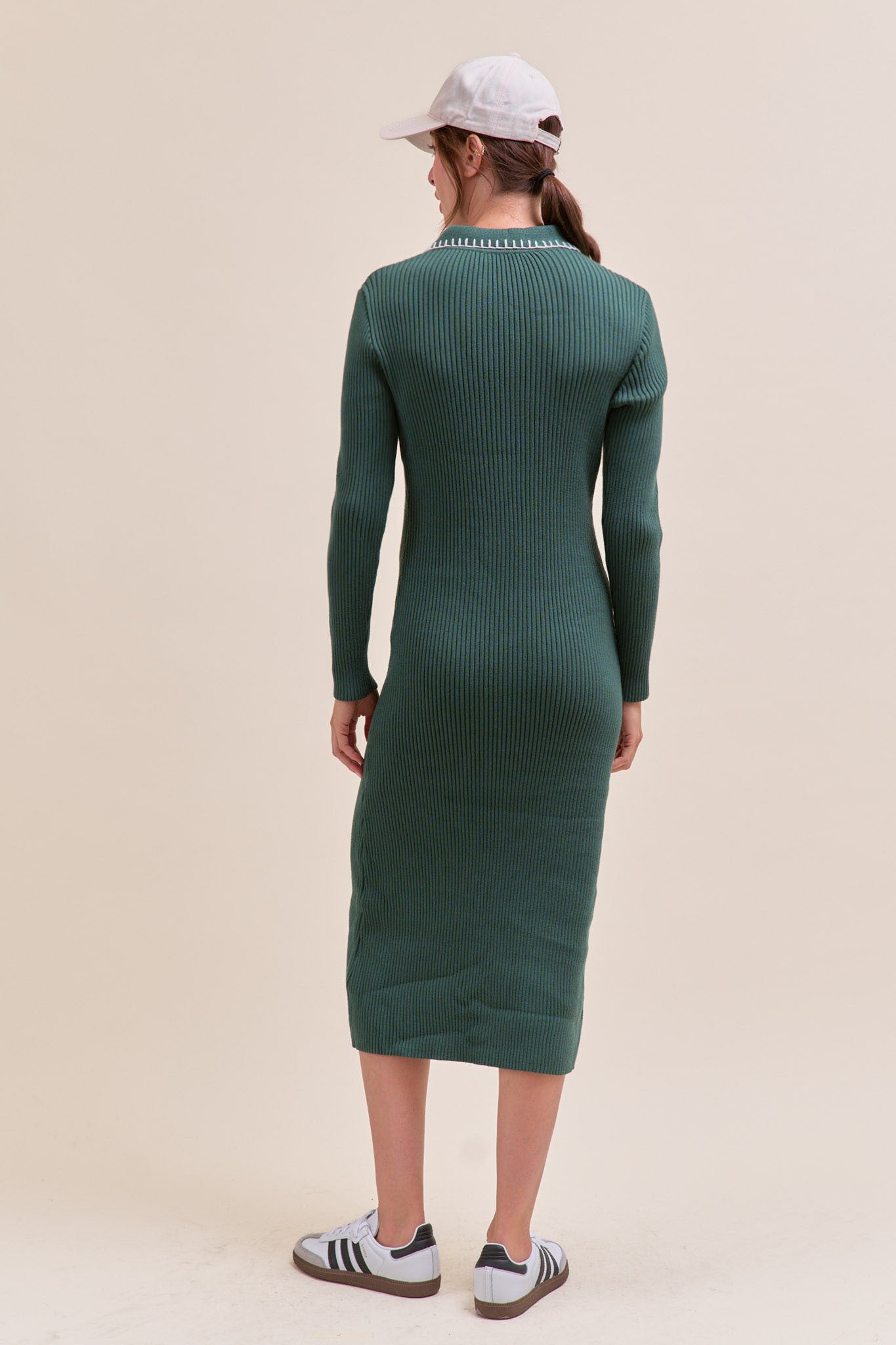 woman wearing a dark green ribbed midi dress with long sleeves, viewed from the back, paired with a white cap and sneakers