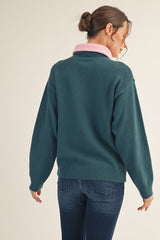 woman wearing a teal oversized sweater with a pink collar and back turned to the camera, paired with dark jeans