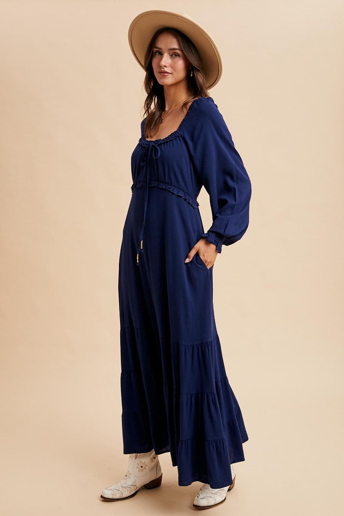 woman wearing a navy blue long sleeve maxi dress with a tiered skirt and a tie detail at the neckline, accessorized with a hat