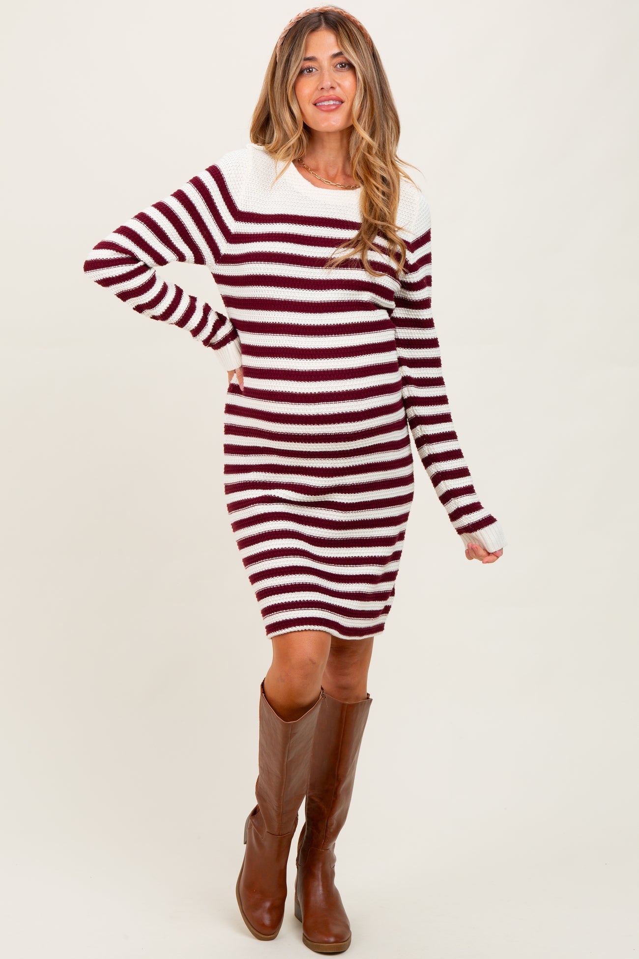 woman wearing a burgundy and white striped maternity sweater dress with long sleeves and standing in knee-high brown boots