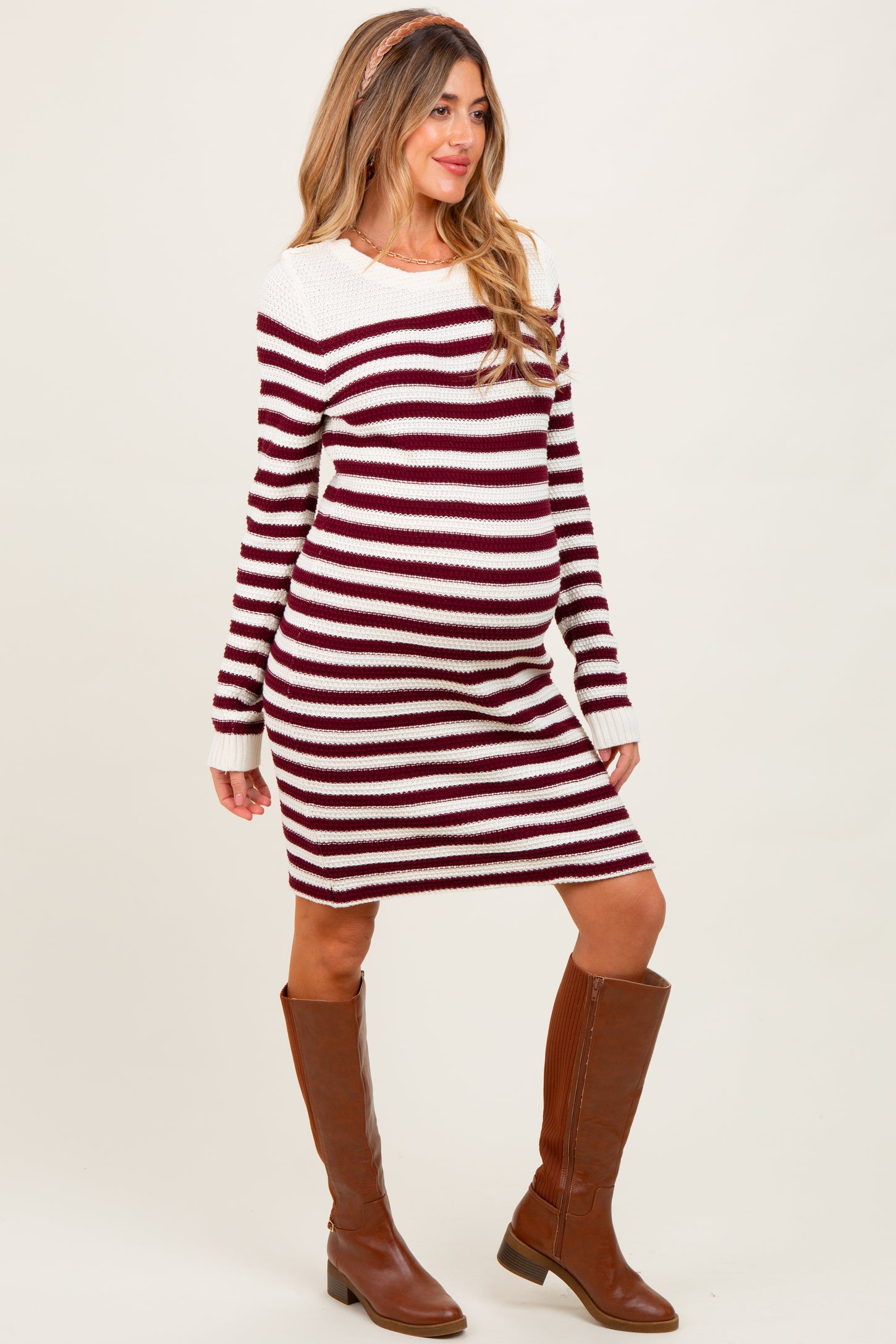 woman wearing a maroon and cream striped maternity sweater dress with a baby bump, paired with brown knee-high boots