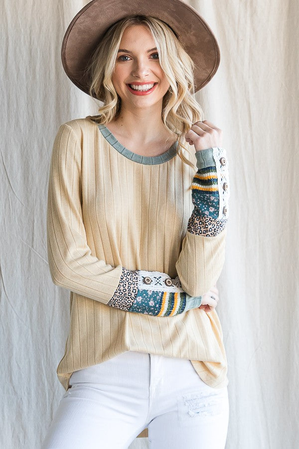 woman wearing a light yellow ribbed long sleeve top with patterned sleeves and a round neckline, smiling while holding her hat
