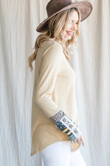 woman wearing a light beige ribbed top with patterned long sleeves and a wide-brimmed hat, smiling against a neutral backdrop