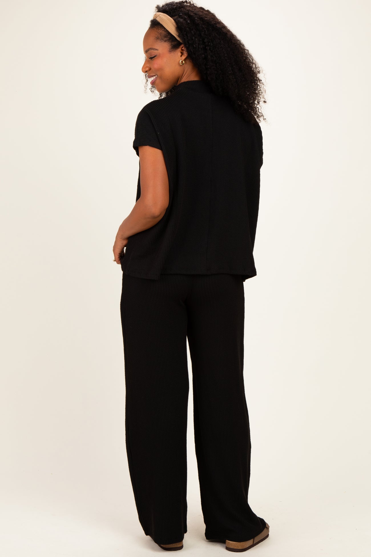 woman wearing a black short sleeve top and matching wide-leg pants, standing sideways with curly hair and a headband