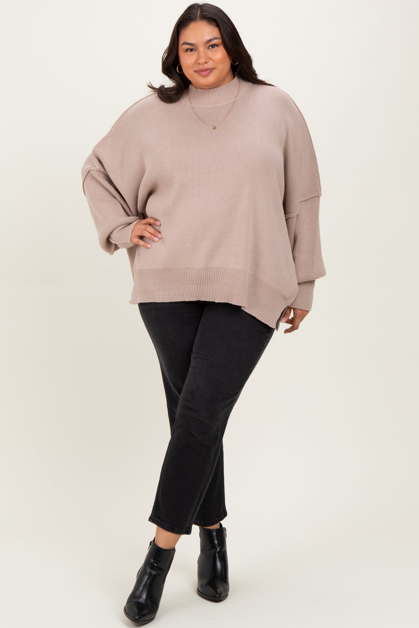 woman wearing a beige oversized knit sweater with a high neckline and black slim-fit jeans, standing confidently