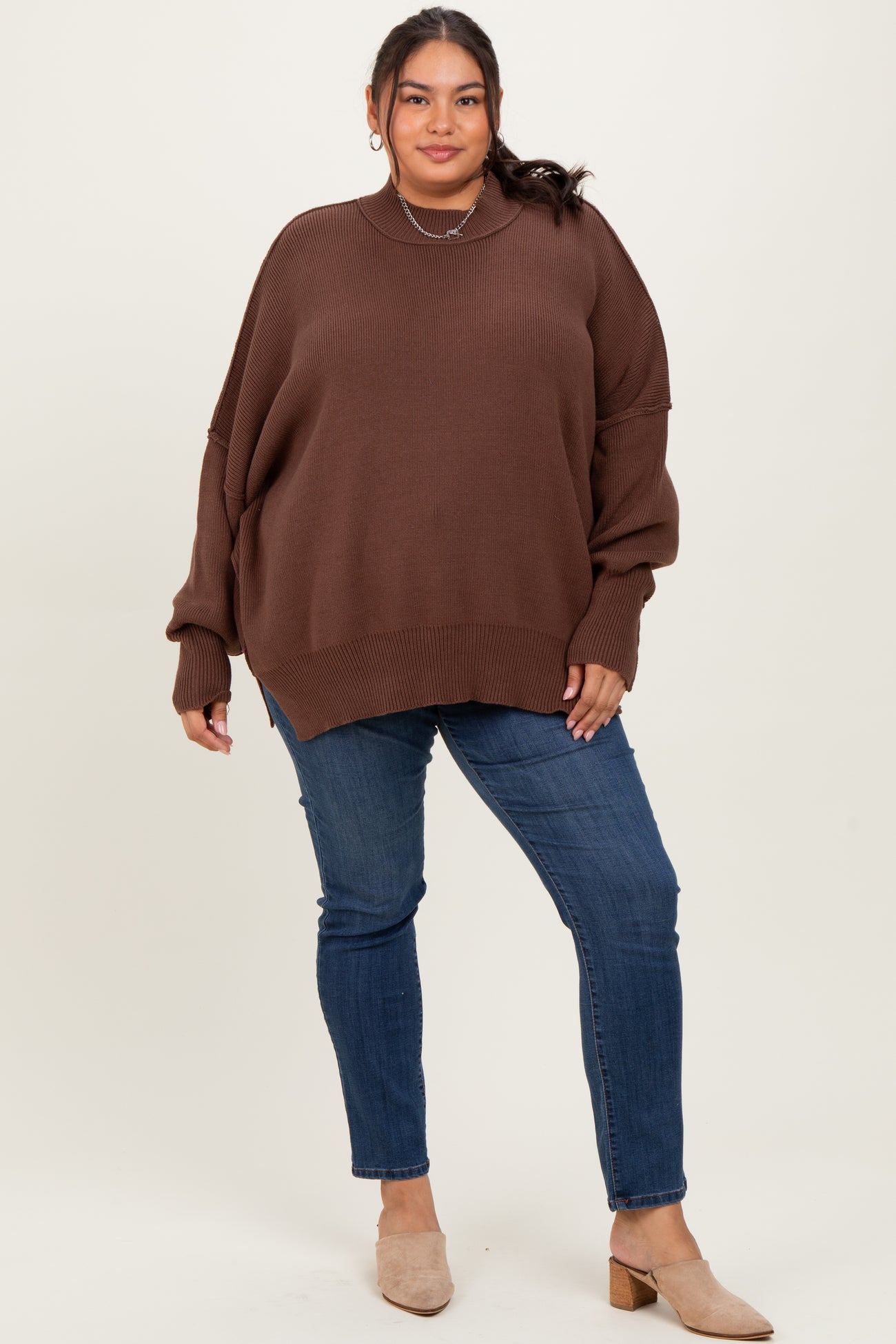 woman wearing a brown oversized sweater and blue skinny jeans, standing against a neutral background