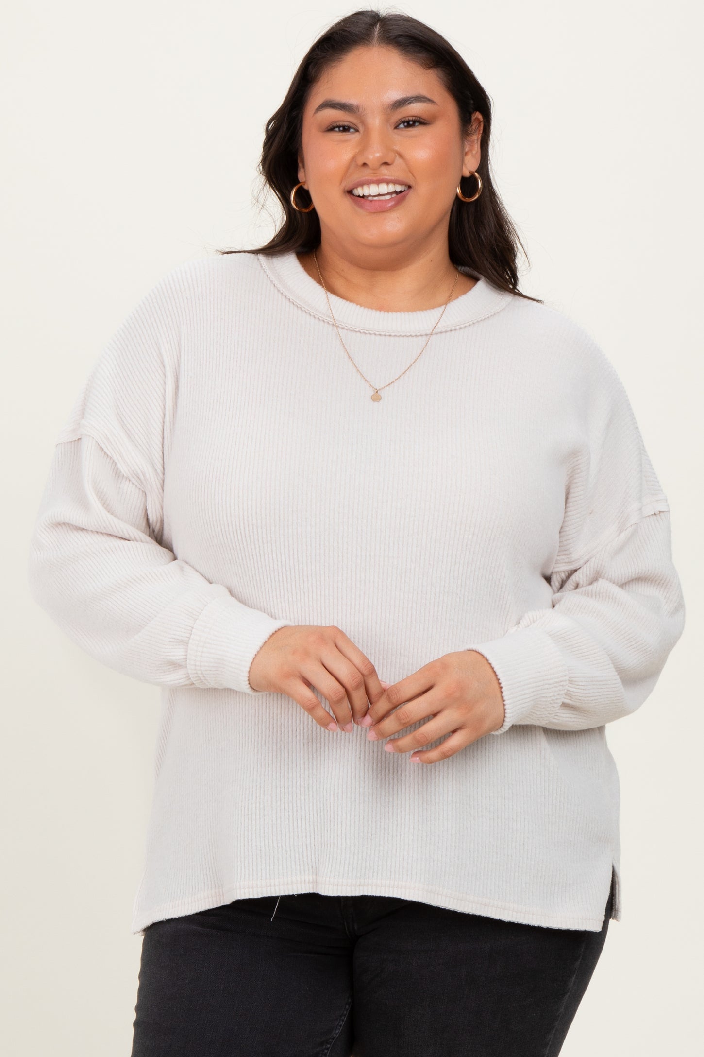 woman wearing a light gray ribbed long sleeve plus size sweater with a relaxed fit and side slits, smiling at the camera