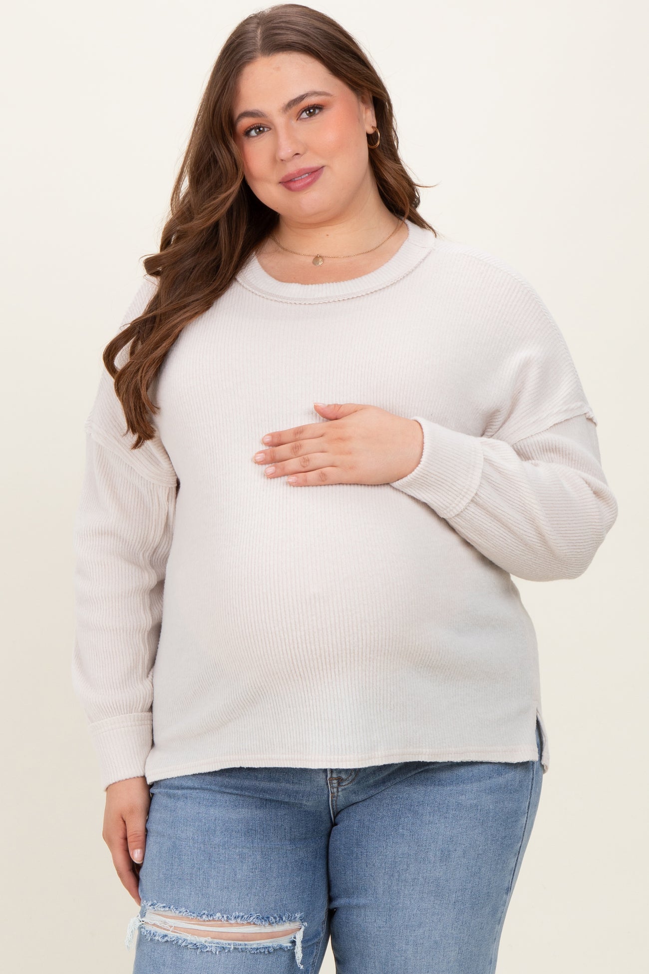 woman wearing a light beige ribbed maternity sweater and holding her baby bump, paired with ripped jeans