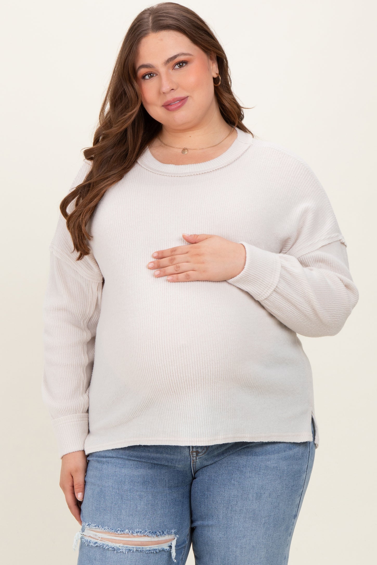 woman wearing a light beige ribbed maternity sweater and holding her baby bump, paired with ripped jeans