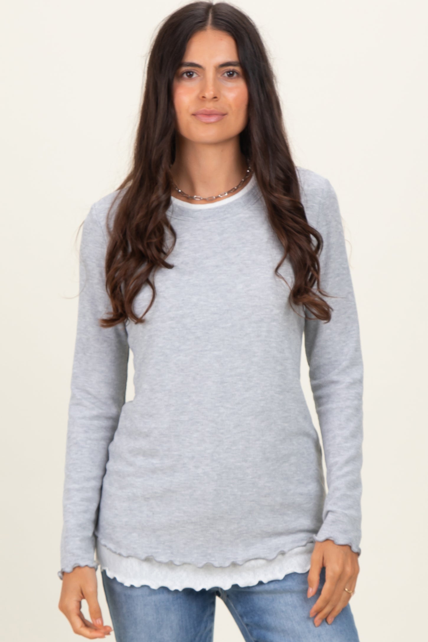 woman wearing a gray long sleeve top with scalloped hem layered over a white shirt and styled with casual jeans
