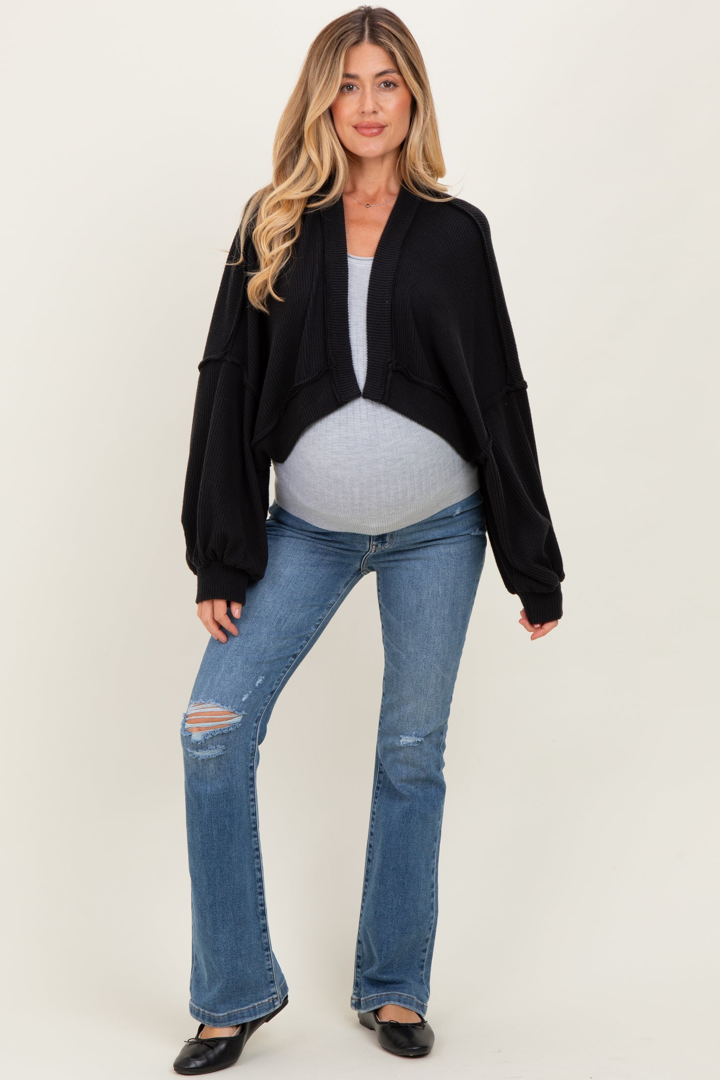 woman wearing a black cropped cardigan over a gray maternity top and flared jeans, showing her baby bump