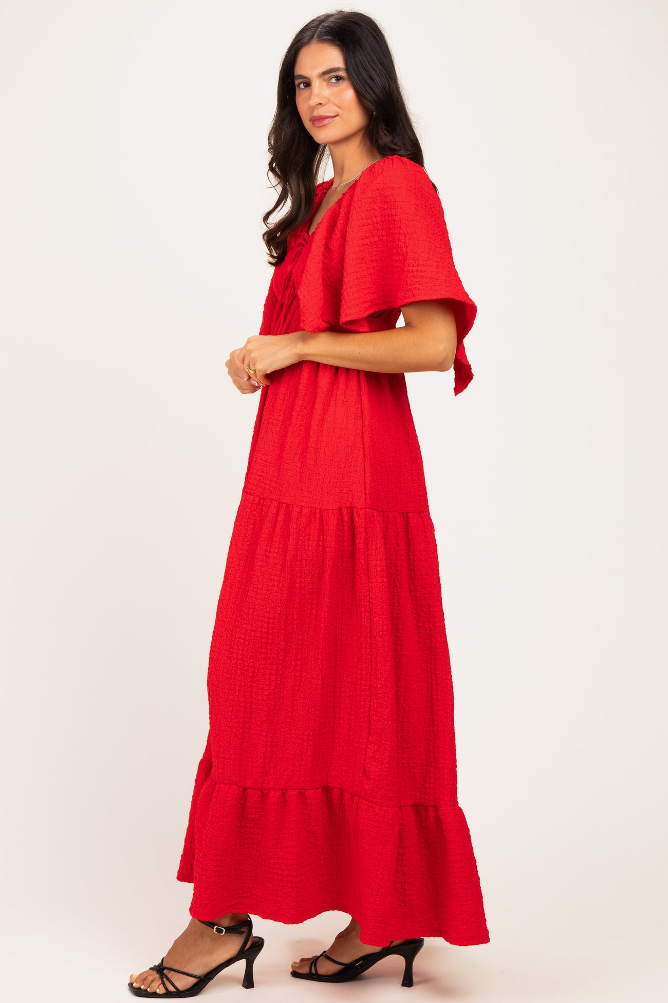 woman wearing a red tiered maxi dress with ruffle sleeves and a bow detail, standing sideways against a plain background