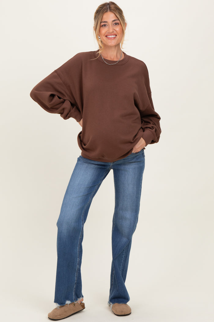 woman wearing a brown oversized maternity sweatshirt and flared jeans, smiling with hands on her hips and a visible baby bump