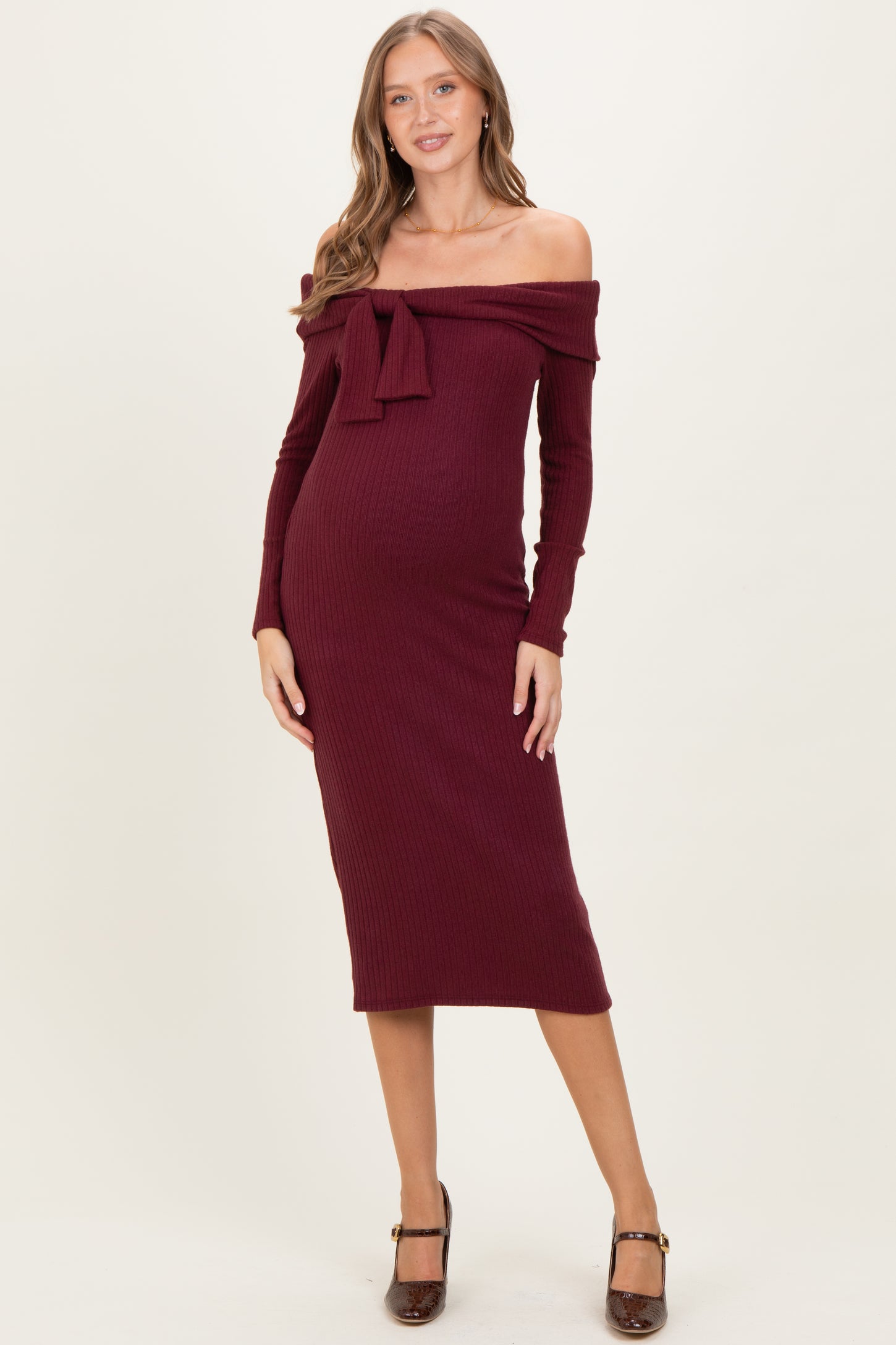 woman wearing a burgundy off-the-shoulder maternity dress with long sleeves and a bow detail, showing her baby bump